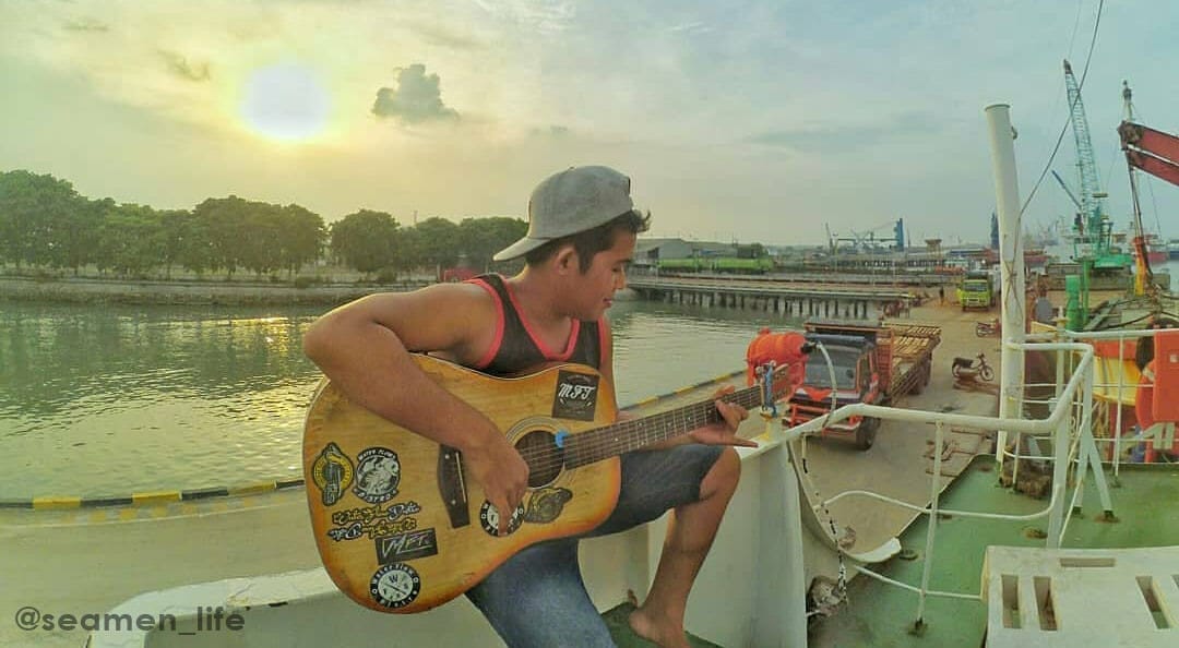 An off duty seafarer playing guitar outside the ship with a view of the port terminal behind him.