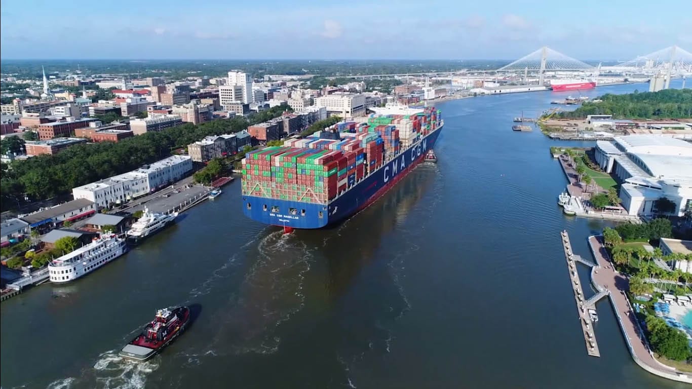 An escort tugboat sailing closely from behind a mega container vessel transiting in a narrow channel.