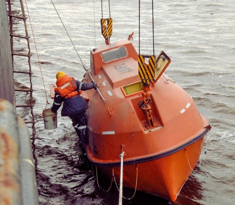 A crew wearing lifejacket holding on to the enclosed lifeboat which is launched on the water.